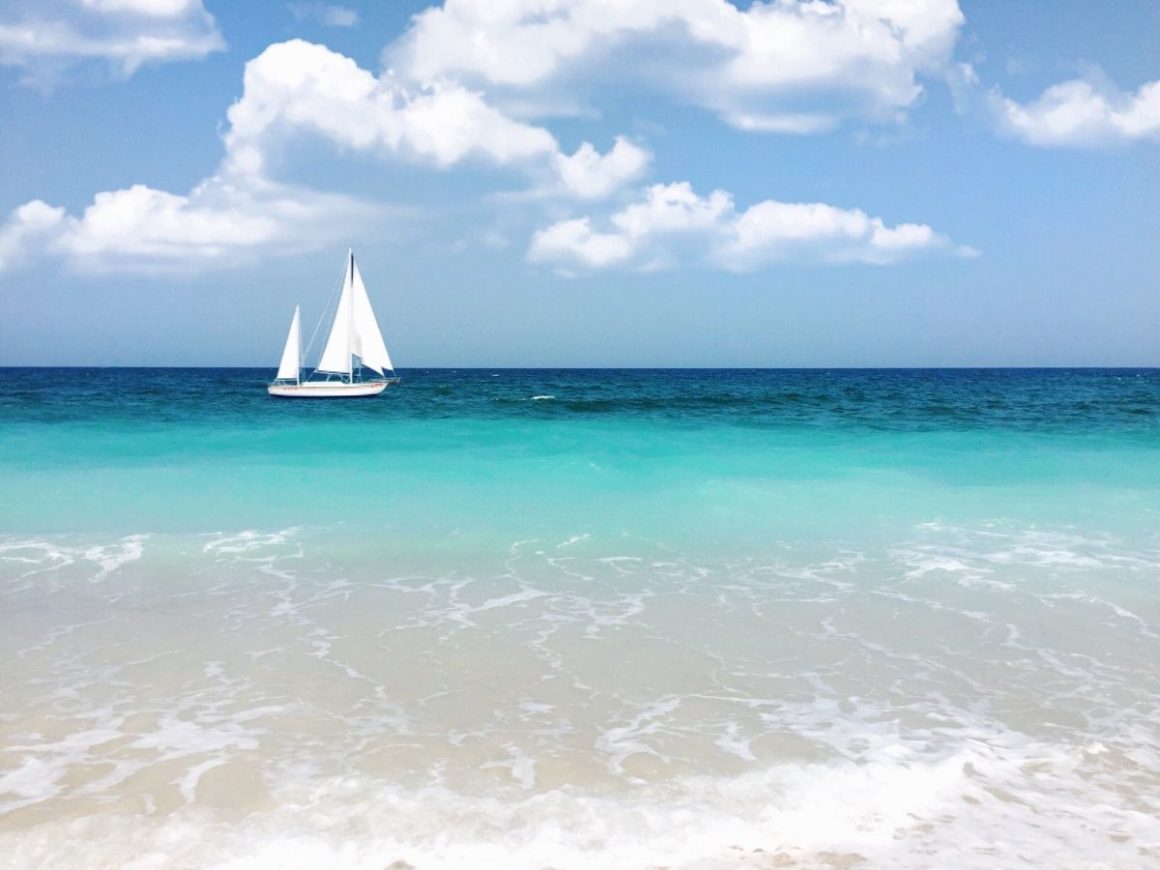 Um veleiro branco desliza pelas águas azuis das Ilhas Turcas e Caicos. A água do mar tem 3 tons, indo do azul escuro, passando pelo azul esverdeado e chegando à praia em um tom claro, misturado com a areia. O céu é azul com nuvens brancas.
