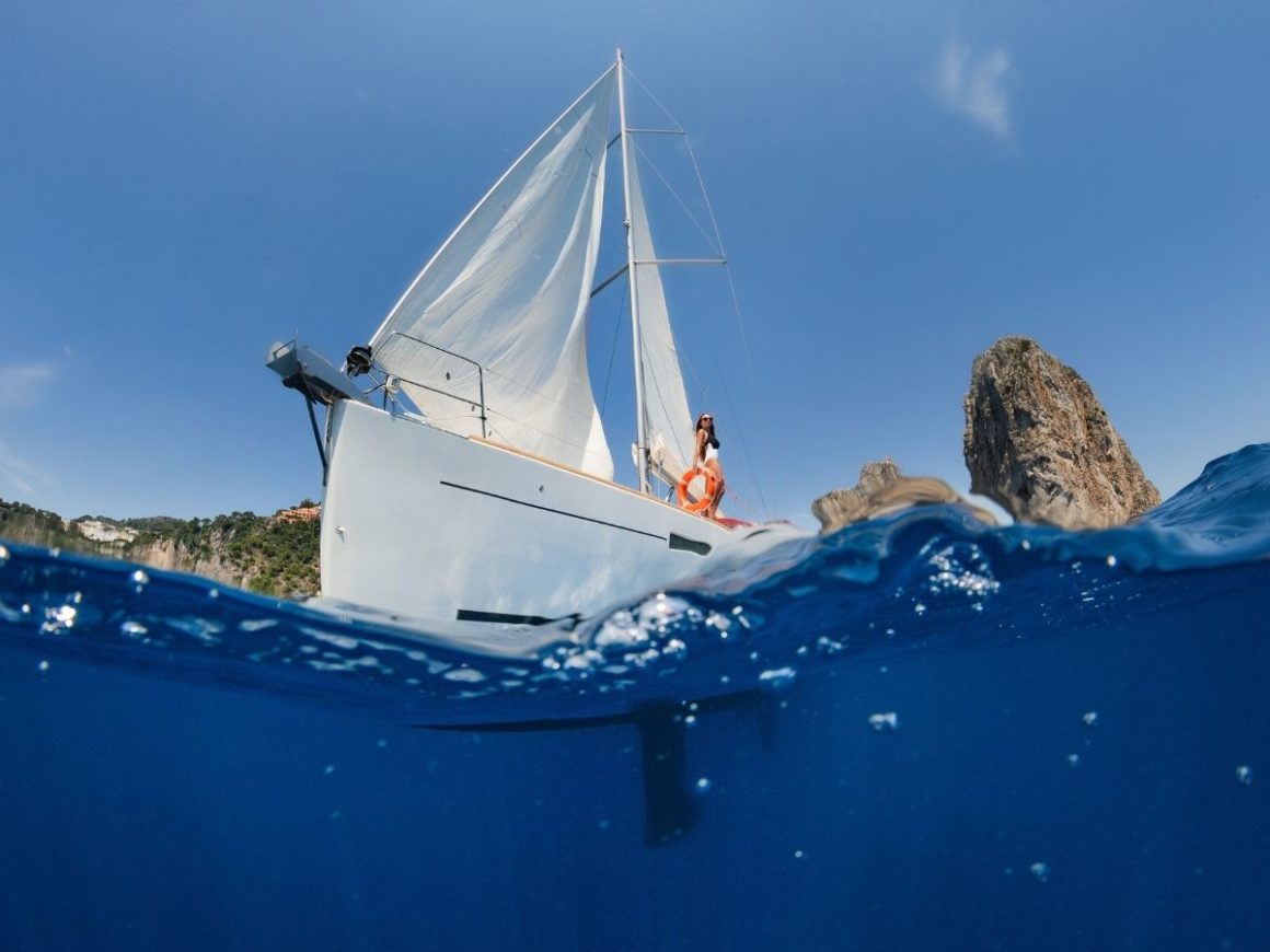 Foto tirada de dentro da água, de cor azul marinho. Acima se vê um veleiro branco e uma mulher vestida de branco. Ao fundo existe uma rocha grande e faz um dia de sol.