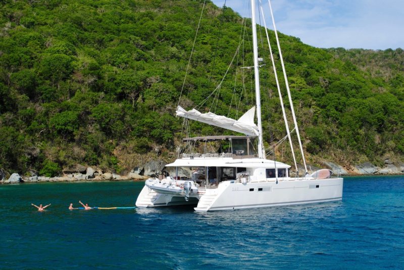 Catamaran anchored on shore with three people swimming at its rear. high outcropping of trees rise from water behind the boat