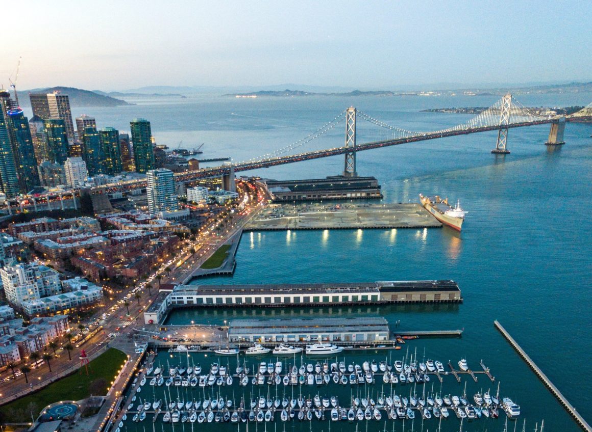 Arial photo of sailboats and yachts parked near the Bay Bridge in San Francisco at dusk.