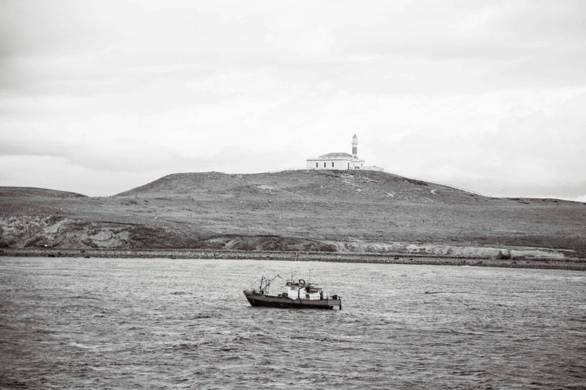 Bateau de pêche naviguant devant le phare de l'île Magdalena en noir et blanc.