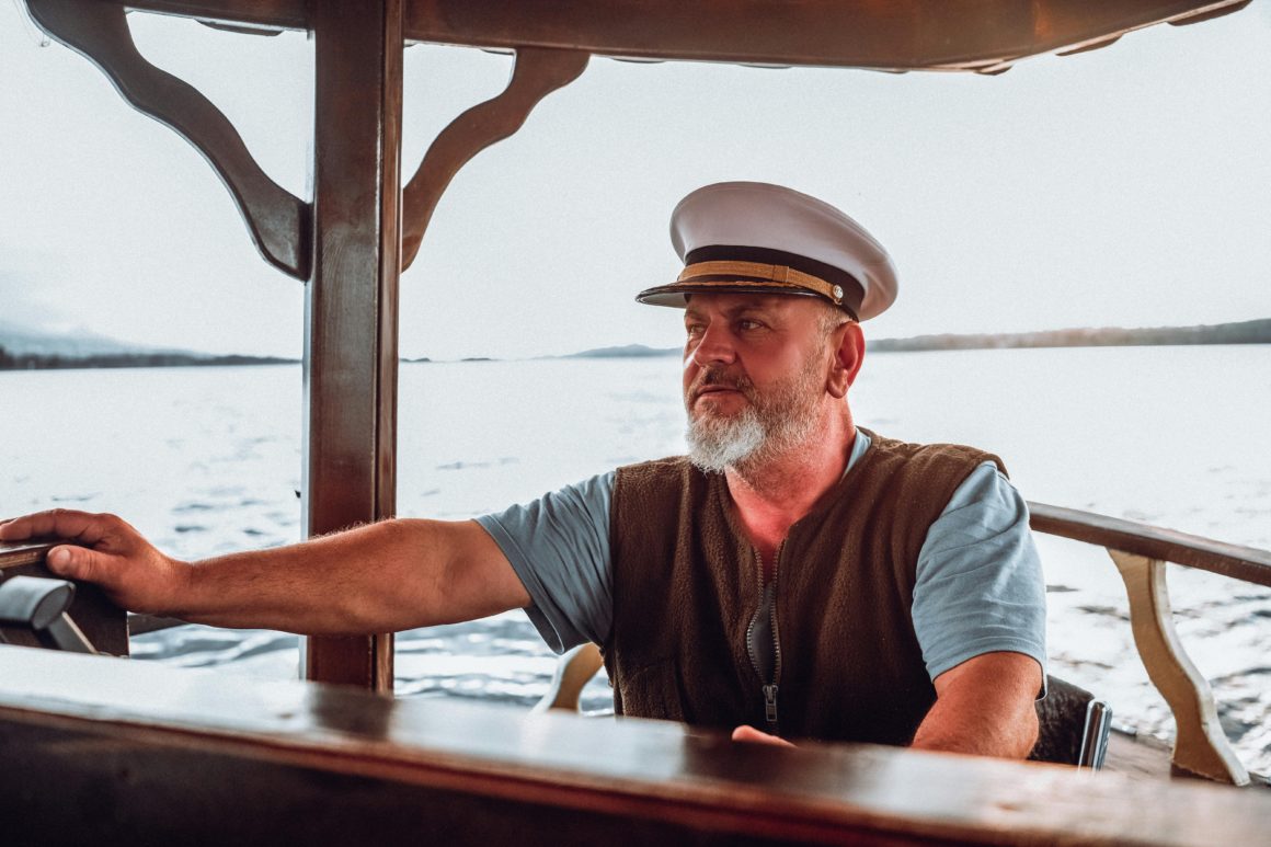 Portrait d'un capitaine expérimenté avec une barbe et une casquette blanche tenant la barre d'un bateau lors d'une navigation calme.