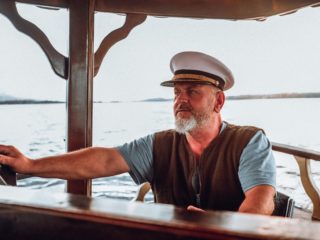 Portrait d'un capitaine expérimenté avec une barbe et une casquette blanche tenant la barre d'un bateau lors d'une navigation calme.