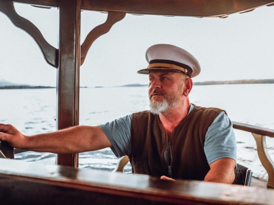 Portrait d'un capitaine expérimenté avec une barbe et une casquette blanche tenant la barre d'un bateau lors d'une navigation calme.