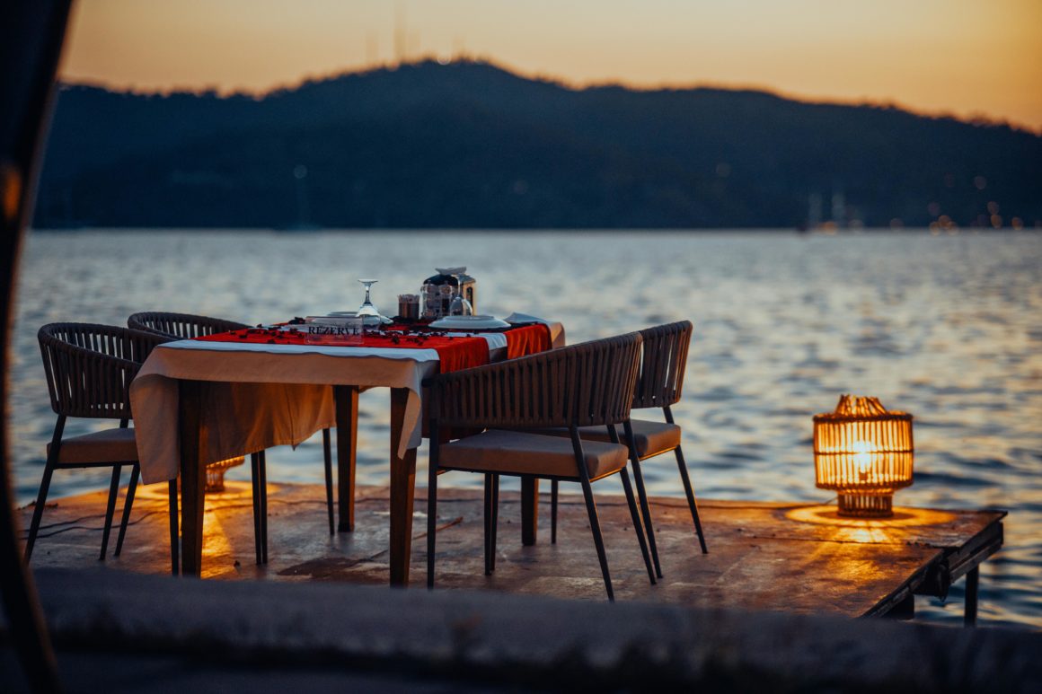 Table dressée pour un dîner romantique sur un ponton en bois au bord de l'eau au crépuscule, avec une lanterne allumée.